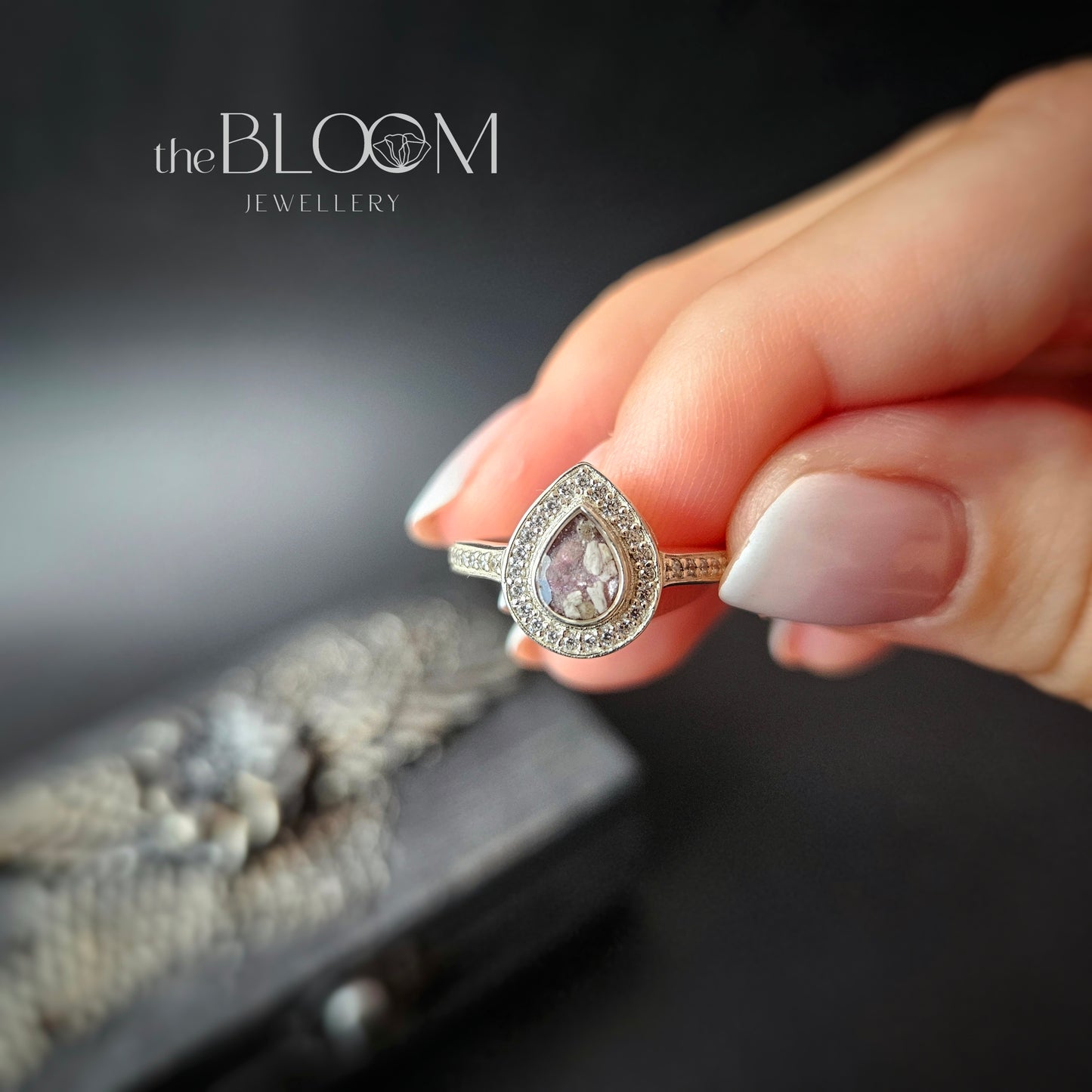 A hand holding a teardrop-style ring with a central cubic zirconia surrounded by smaller clear crystals, set in a sterling silver band.