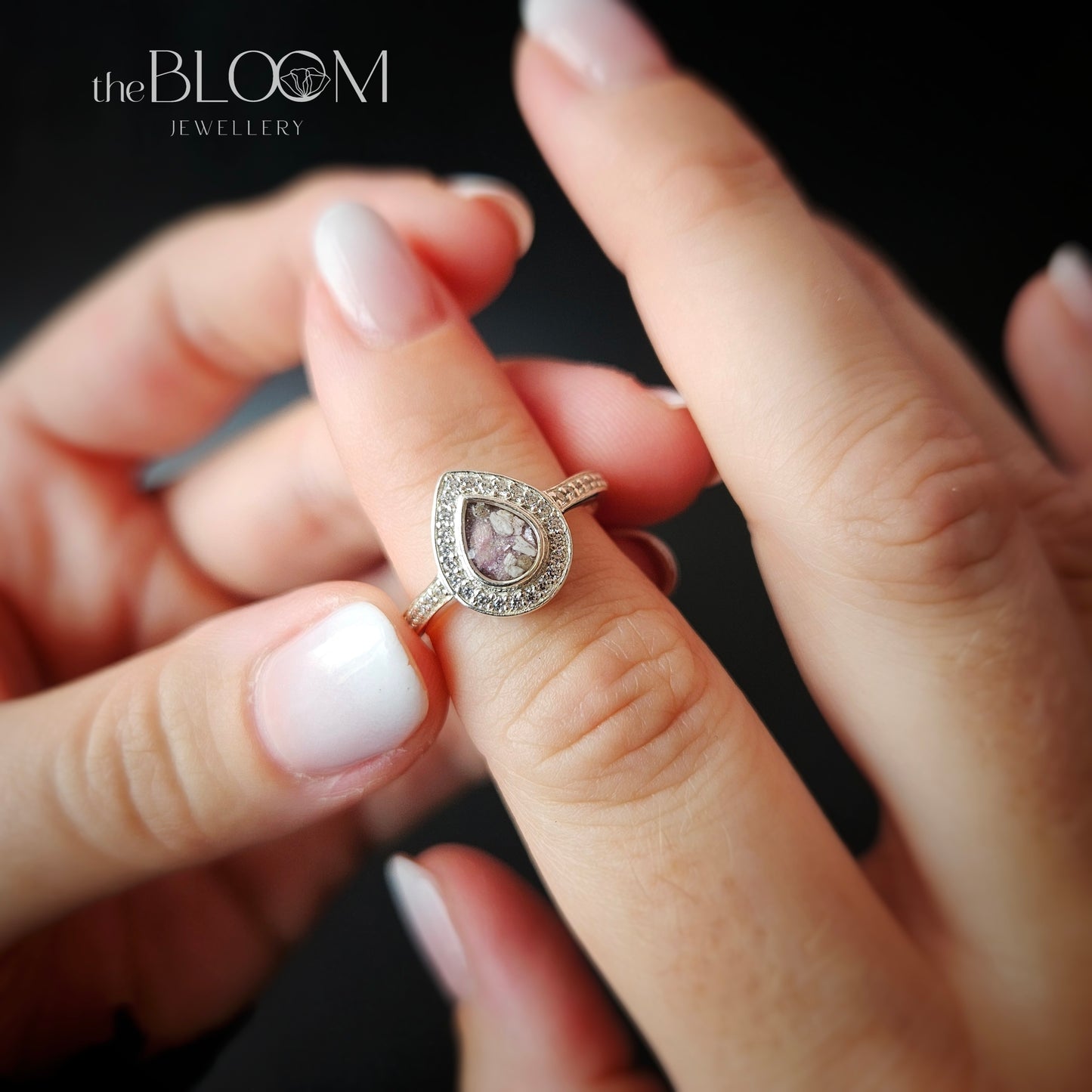A teardrop-shaped silver ring with a central clear gemstone and surrounded by cubic zirconia, held between two fingers.