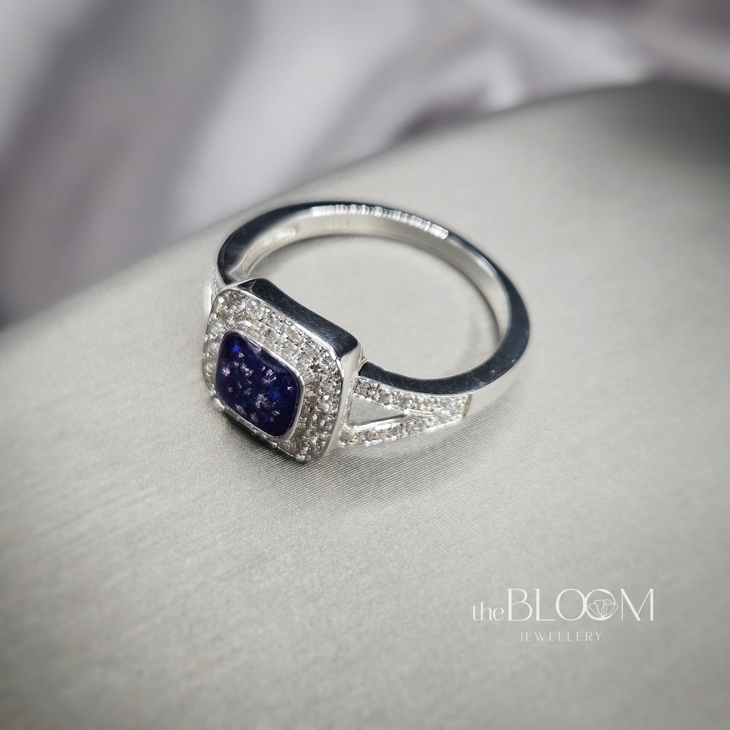 Silver ring with a blue gemstone on a gray surface, branded 'the BLOOM Jewellery'.