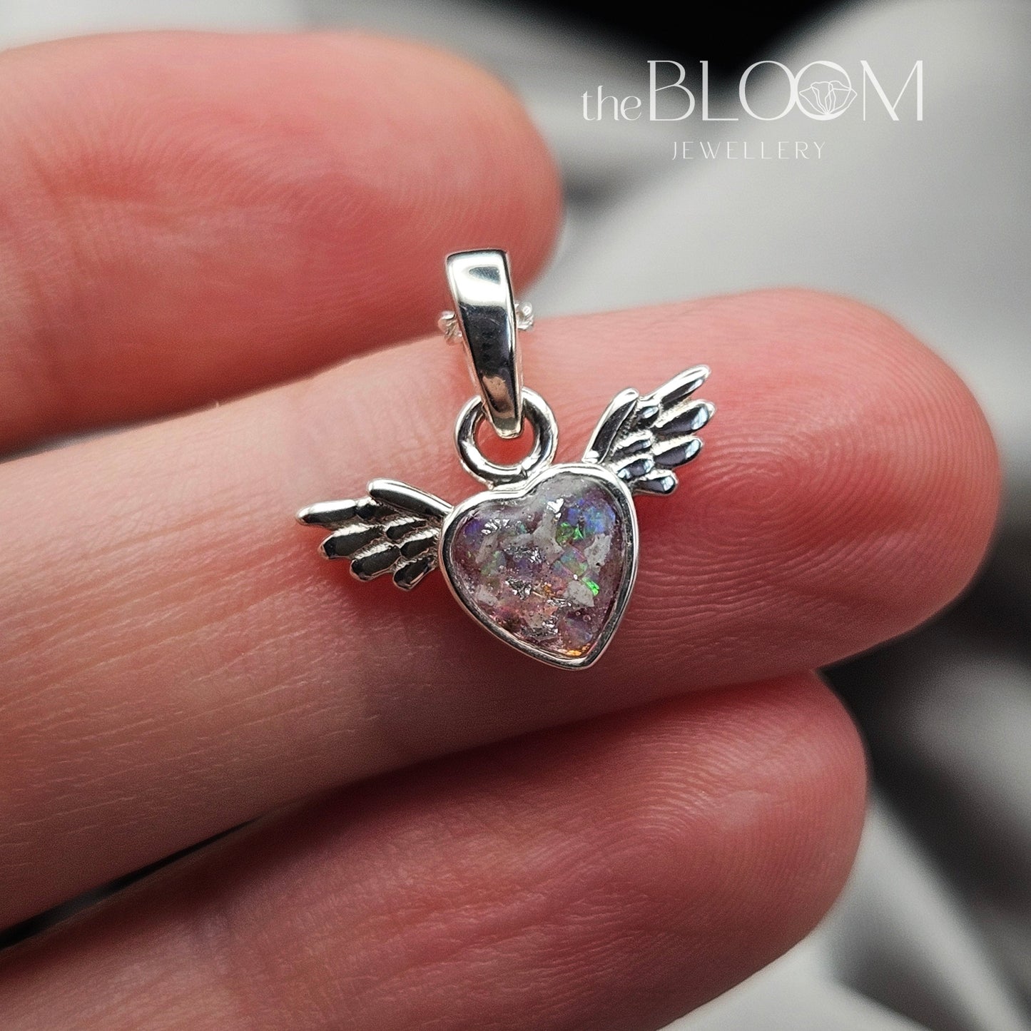 Silver heart-shaped pendant with angel wings held between fingers, brand 'the BLOOM' visible.