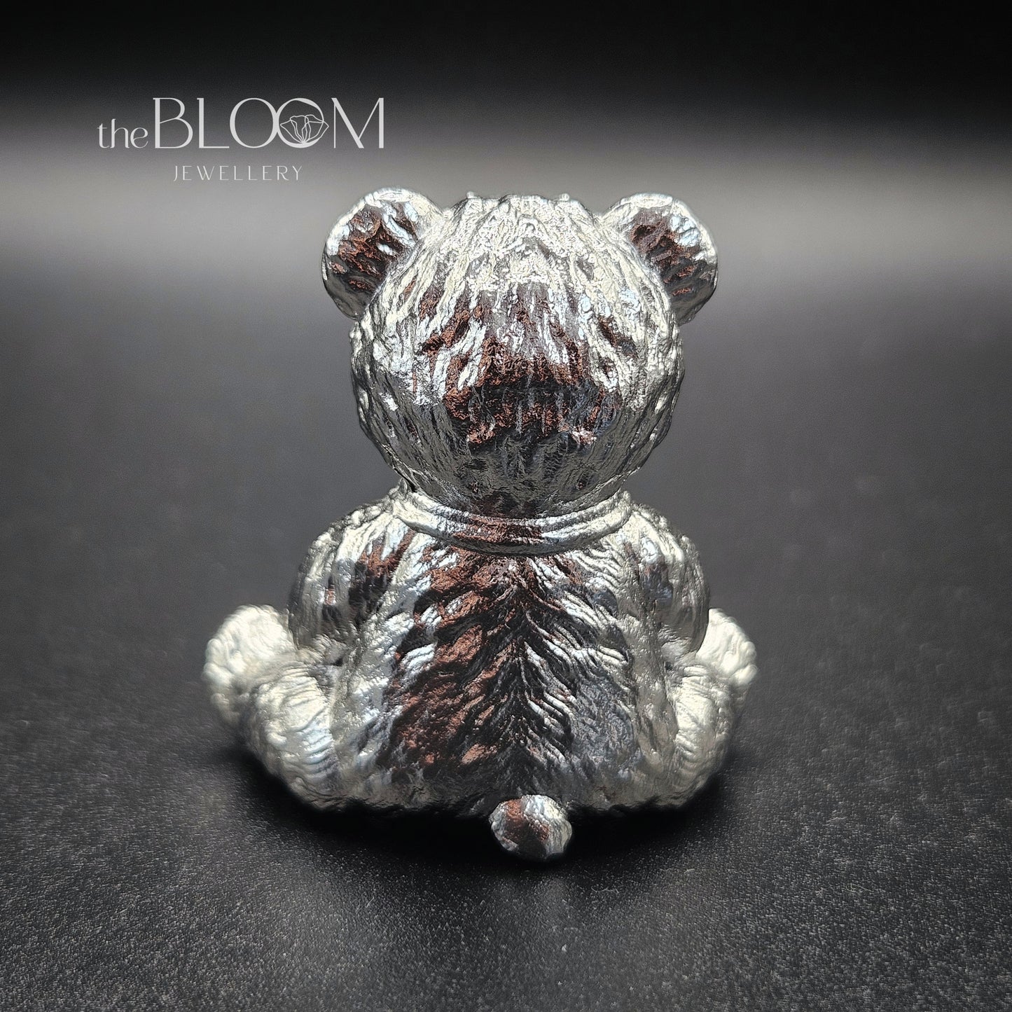Silver teddy bear figurine on a dark background with 'the BLOOM JEWELLERY' branding.