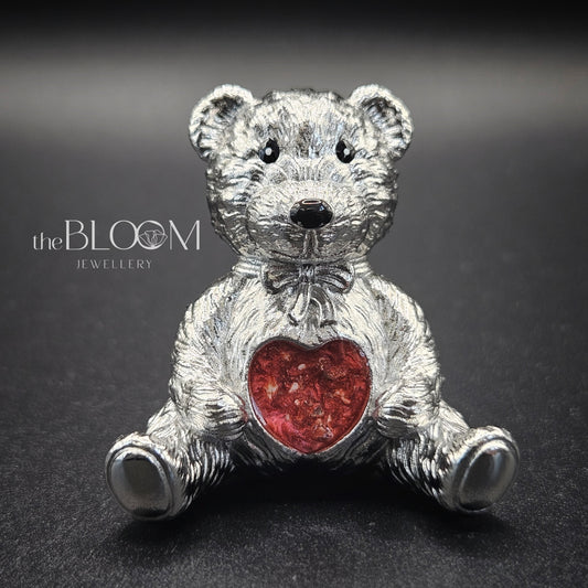 Silver teddy bear with a red heart on a dark background, branded 'the BLOOM JEWELLERY'.