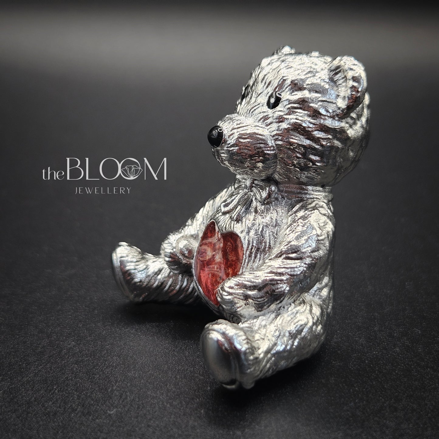 Silver teddy bear figurine holding a red heart on a dark background with 'the BLOOM JEWELLERY' branding.