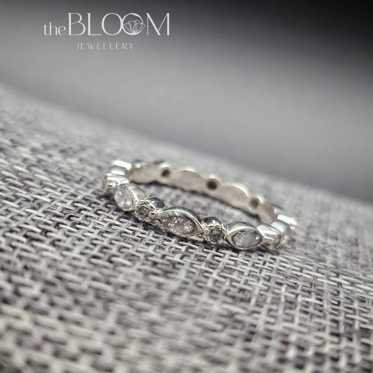 Silver ring with small stones on a textured fabric surface, branded 'the BLOOM Jewellery'.