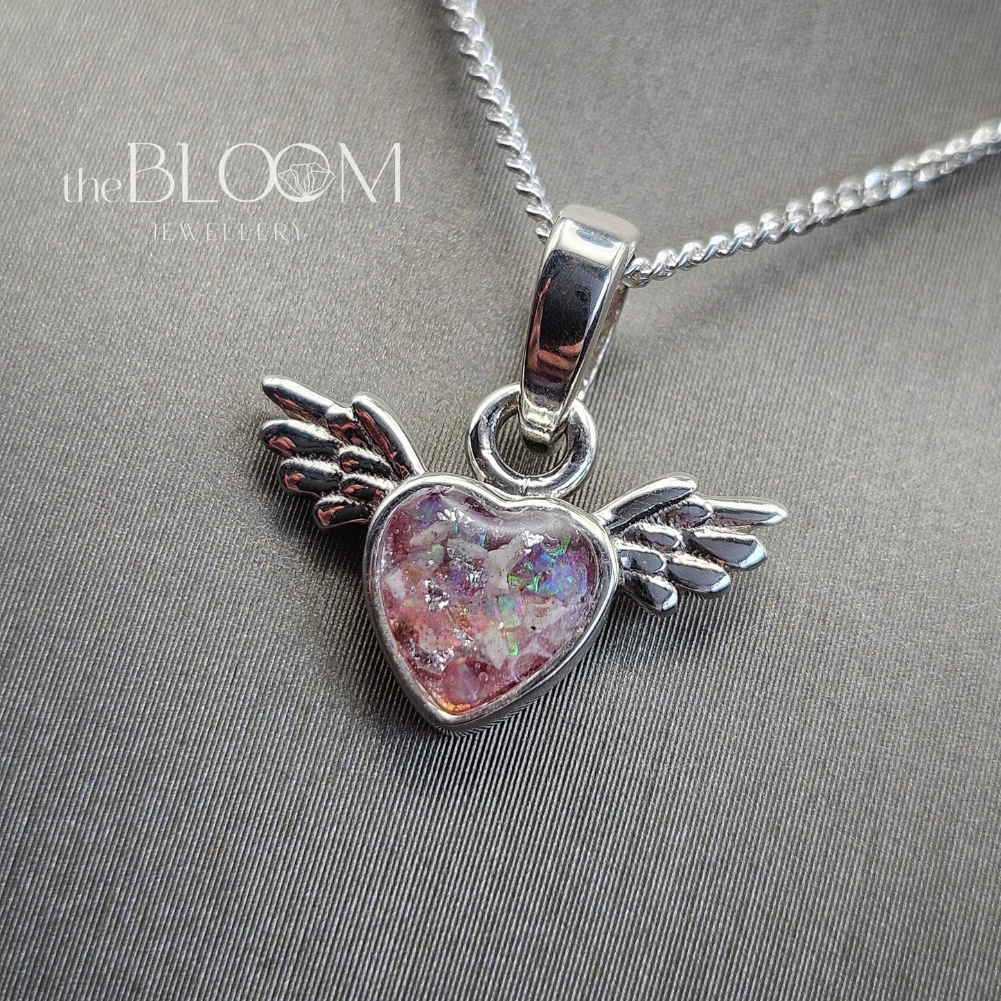 Heart-shaped pendant with wings on a chain against a gray background, branded 'the BLOOM'.