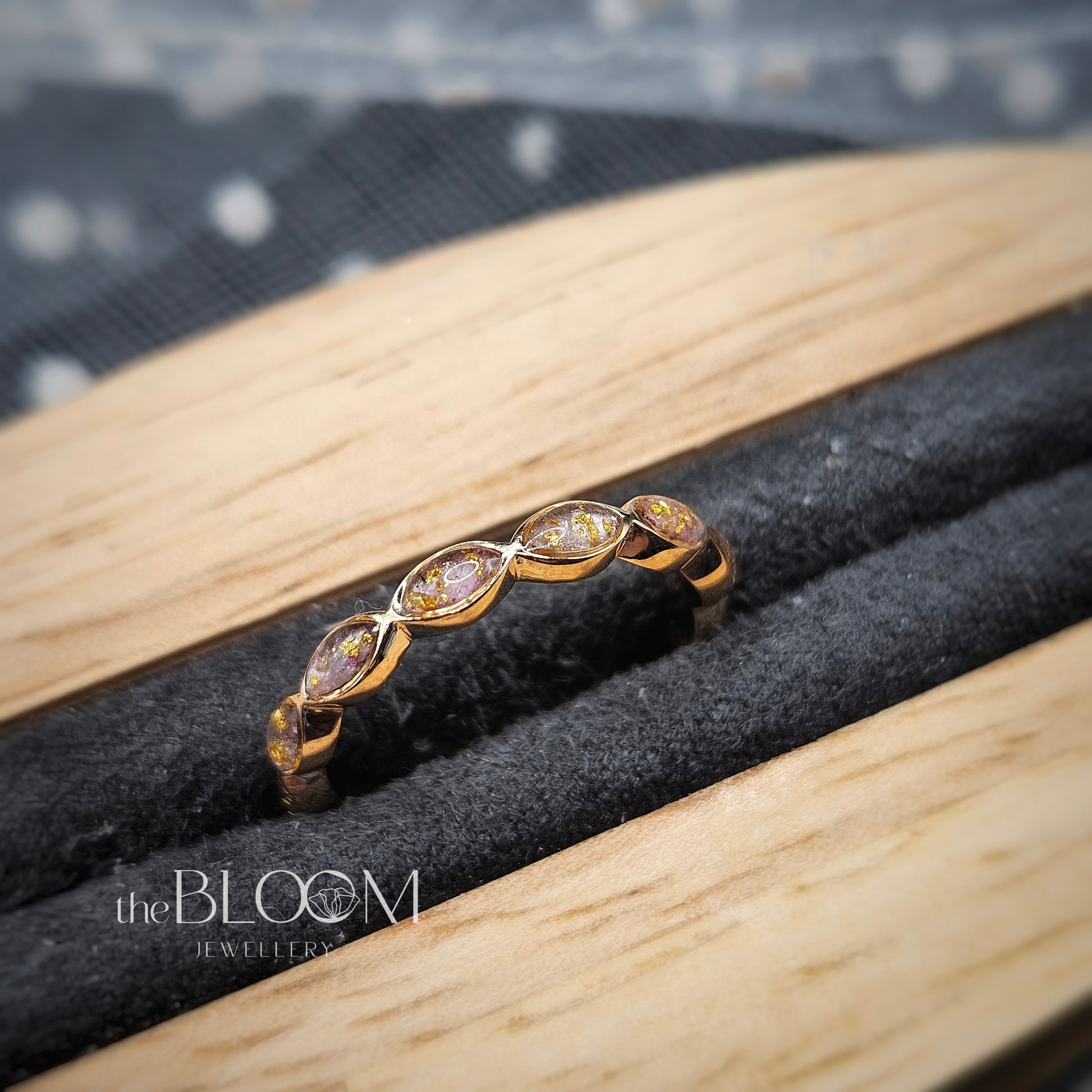 Gold ring with ashes gemstones on a wooden and stone surface, branded 'the BLOOM JEWELLERY'.