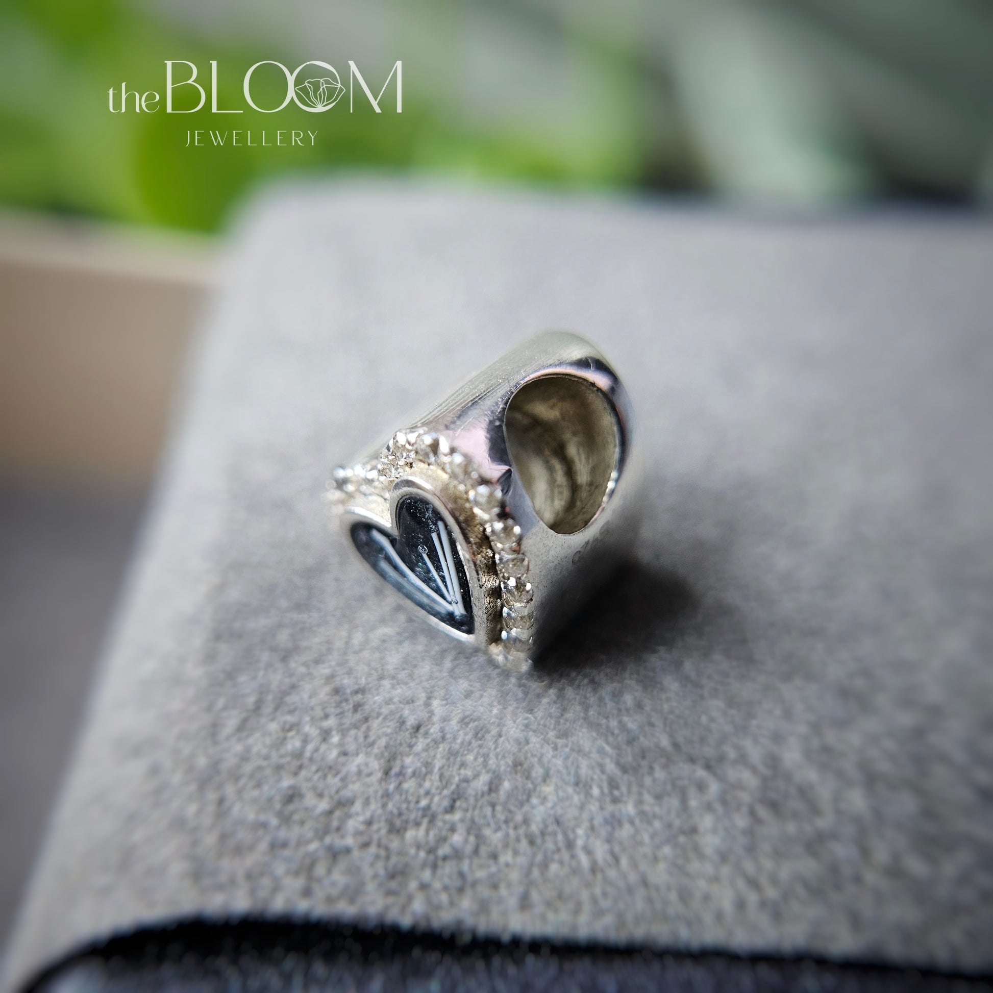 Silver charm with heart design on a gray surface, branded 'the Bloom Jewellery'.
