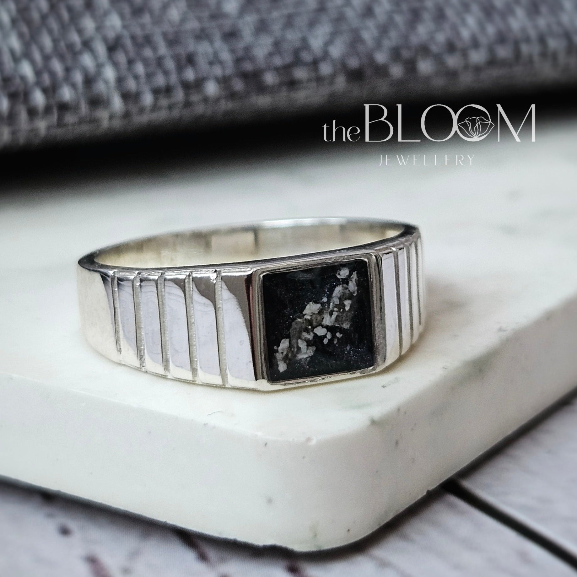 Silver ring with a black stone on a textured surface, branded 'theBLOOM'.