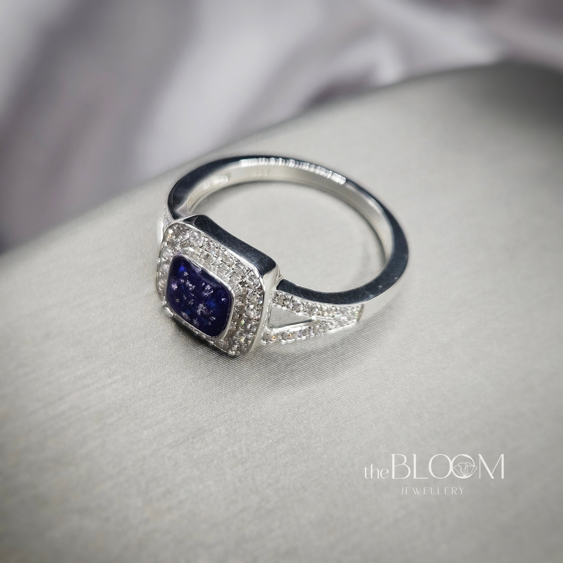 Silver ring with a blue gemstone on a gray surface, branded 'the BLOOM Jewellery'.