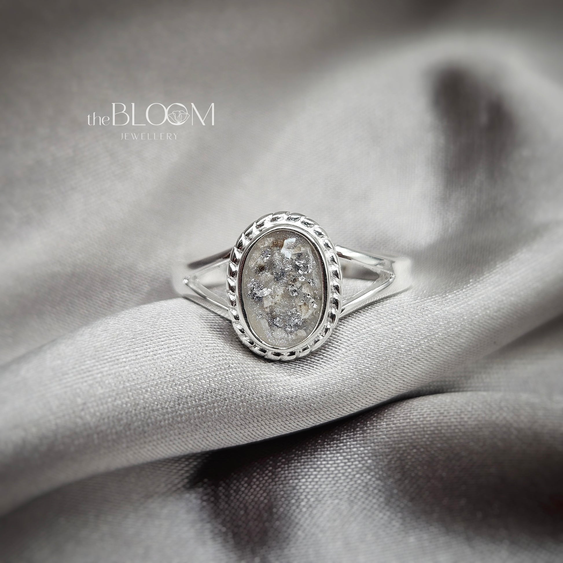 Silver ring with a central stone on a gray fabric background, featuring 'the BLOOM Jewellery' brand.