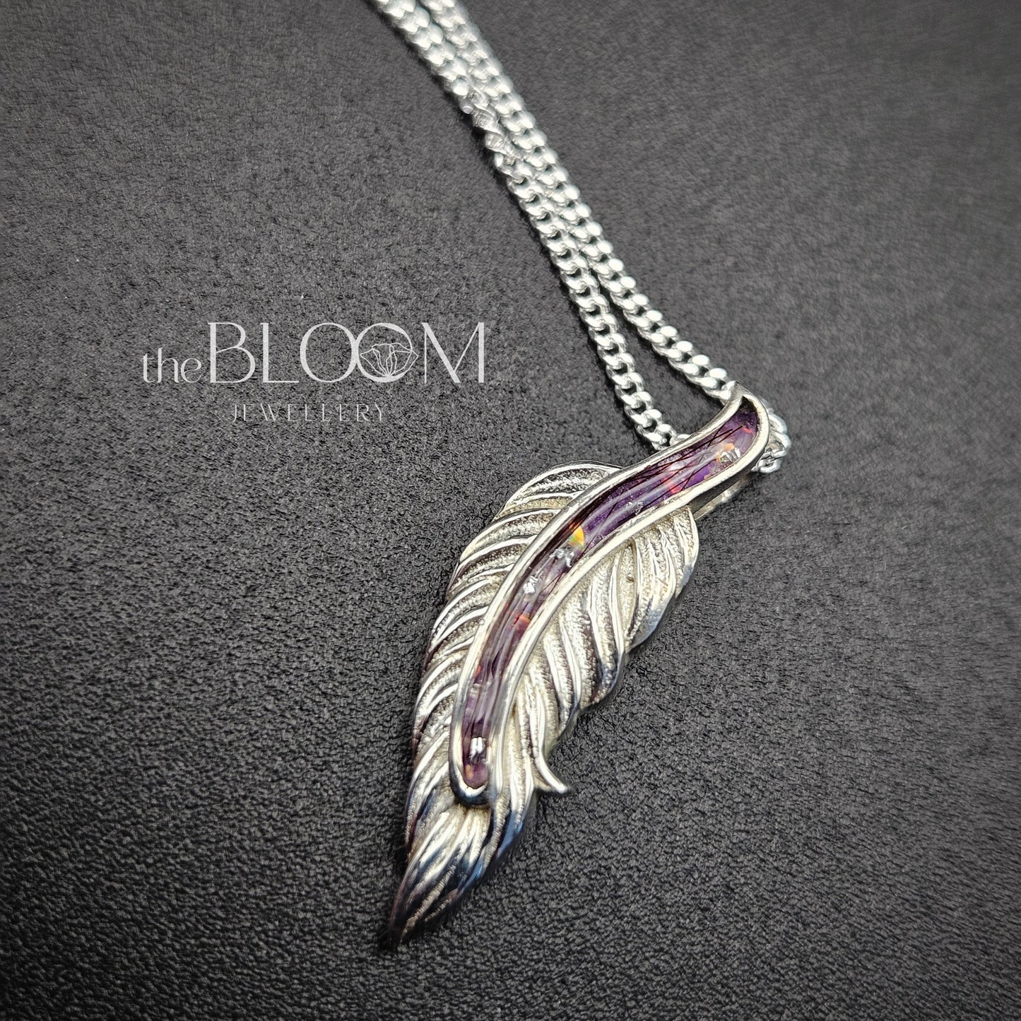Silver feather-shaped pendant on a chain against a dark background with 'the Bloom' brand visible.