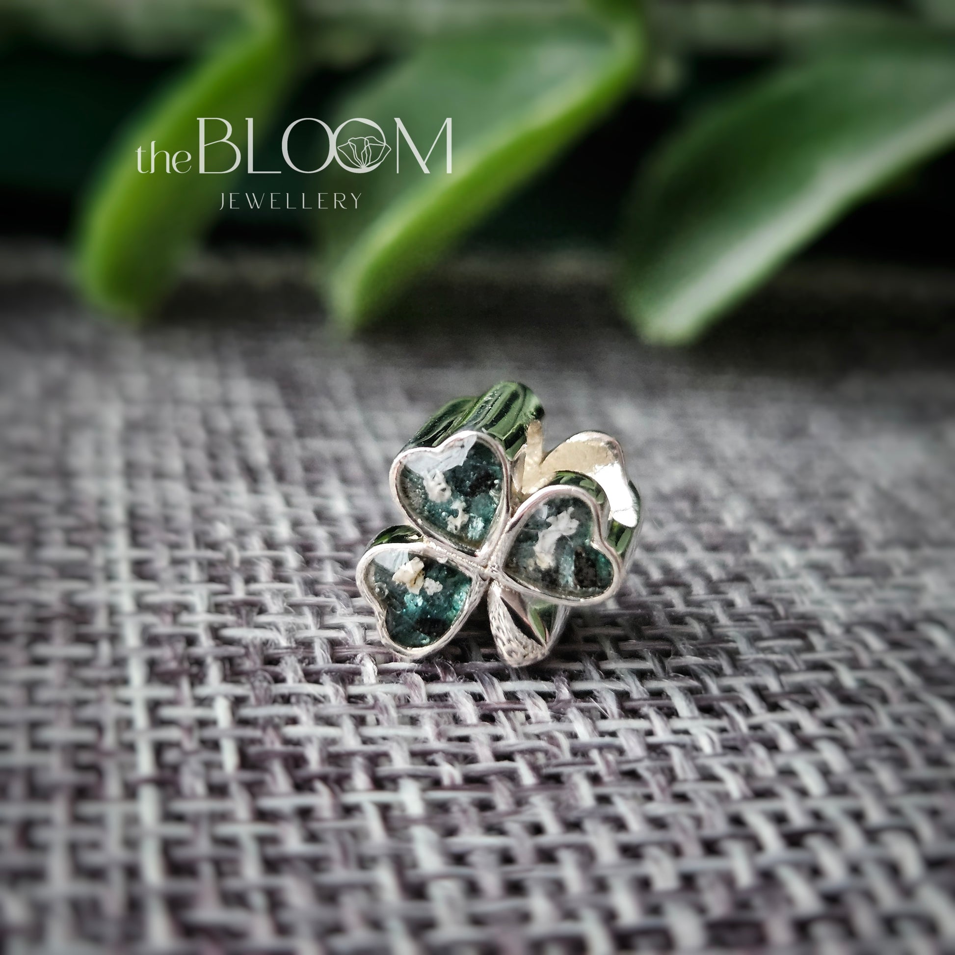Silver four-leaf clover pendant on a textured fabric background with 'the BLOOM JEWELLERY' branding.