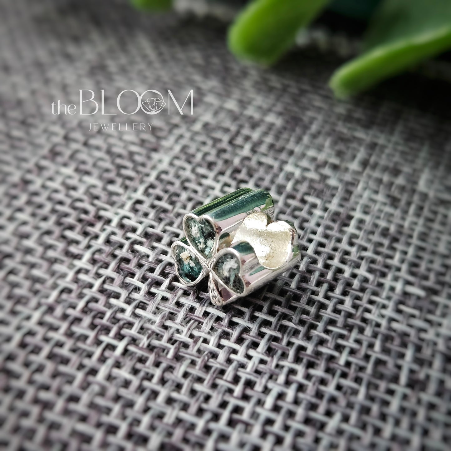 Silver jewelry piece with green stones on a textured fabric background, branded 'the BLOOM Jewellery'.