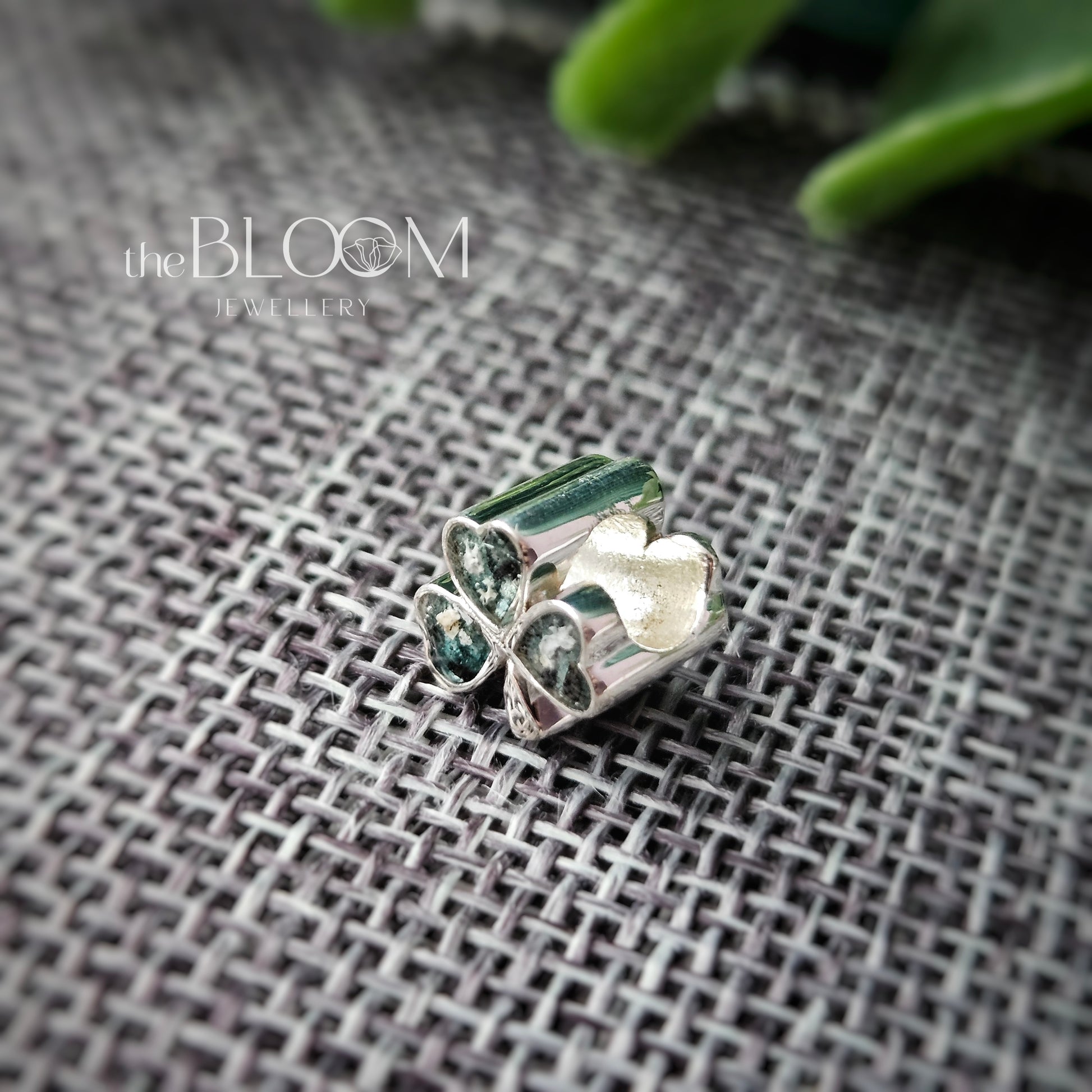 Silver jewelry piece with green stones on a textured fabric background, branded 'the BLOOM Jewellery'.