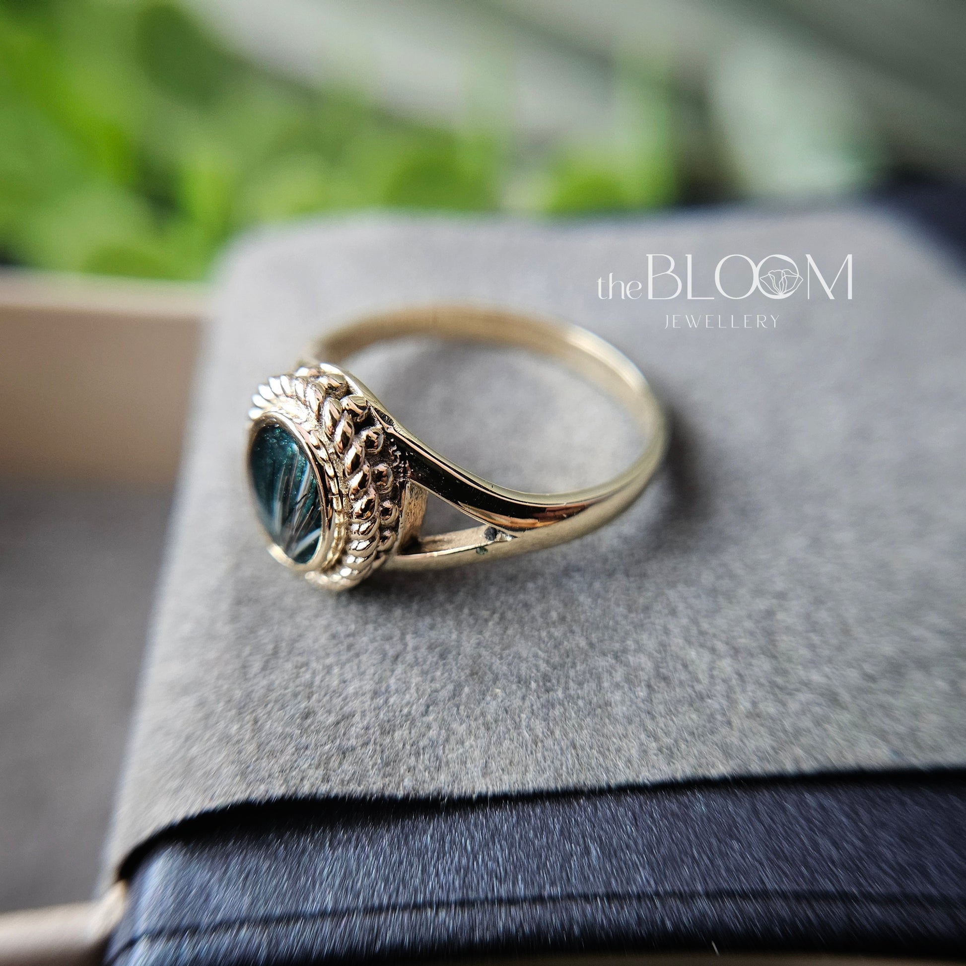 Gold ring with blue stone on a gray surface, branded 'the BLOOM Jewellery'.