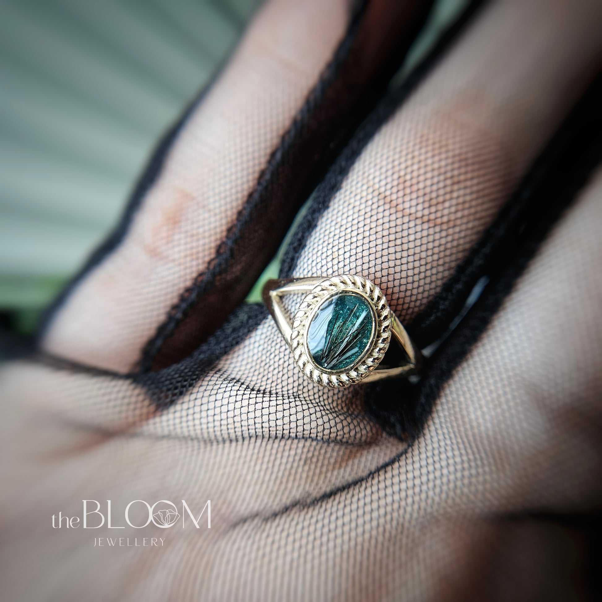 Gold ring with a green stone on a hand wearing black mesh glove, brand 'the BLOOM JEWELLERY' visible.