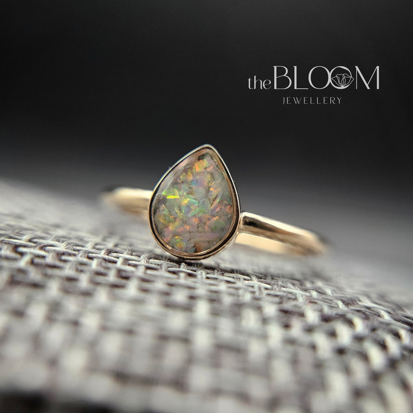 Gold ring with a teardrop-shaped opal stone on a textured surface, branded 'the BLOOM'.