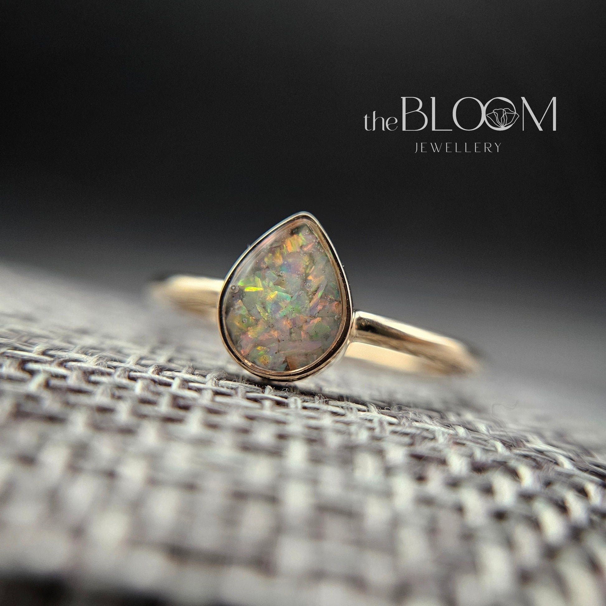 Gold ring with a teardrop-shaped opal stone on a textured surface, branded 'the BLOOM'.