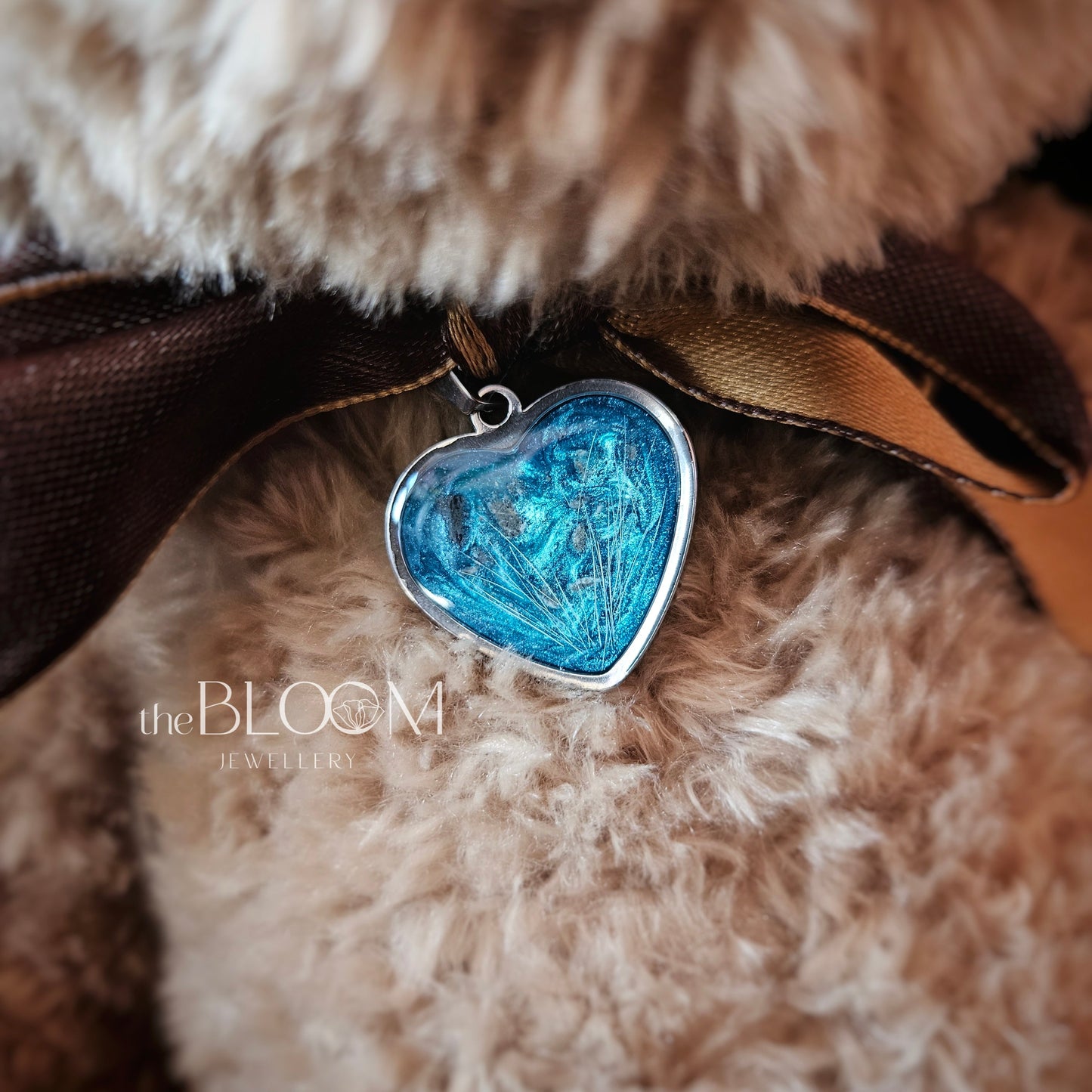 Blue heart-shaped pendant on a brown ribbon against a fluffy beige background with 'the BLOOM' brand visible.