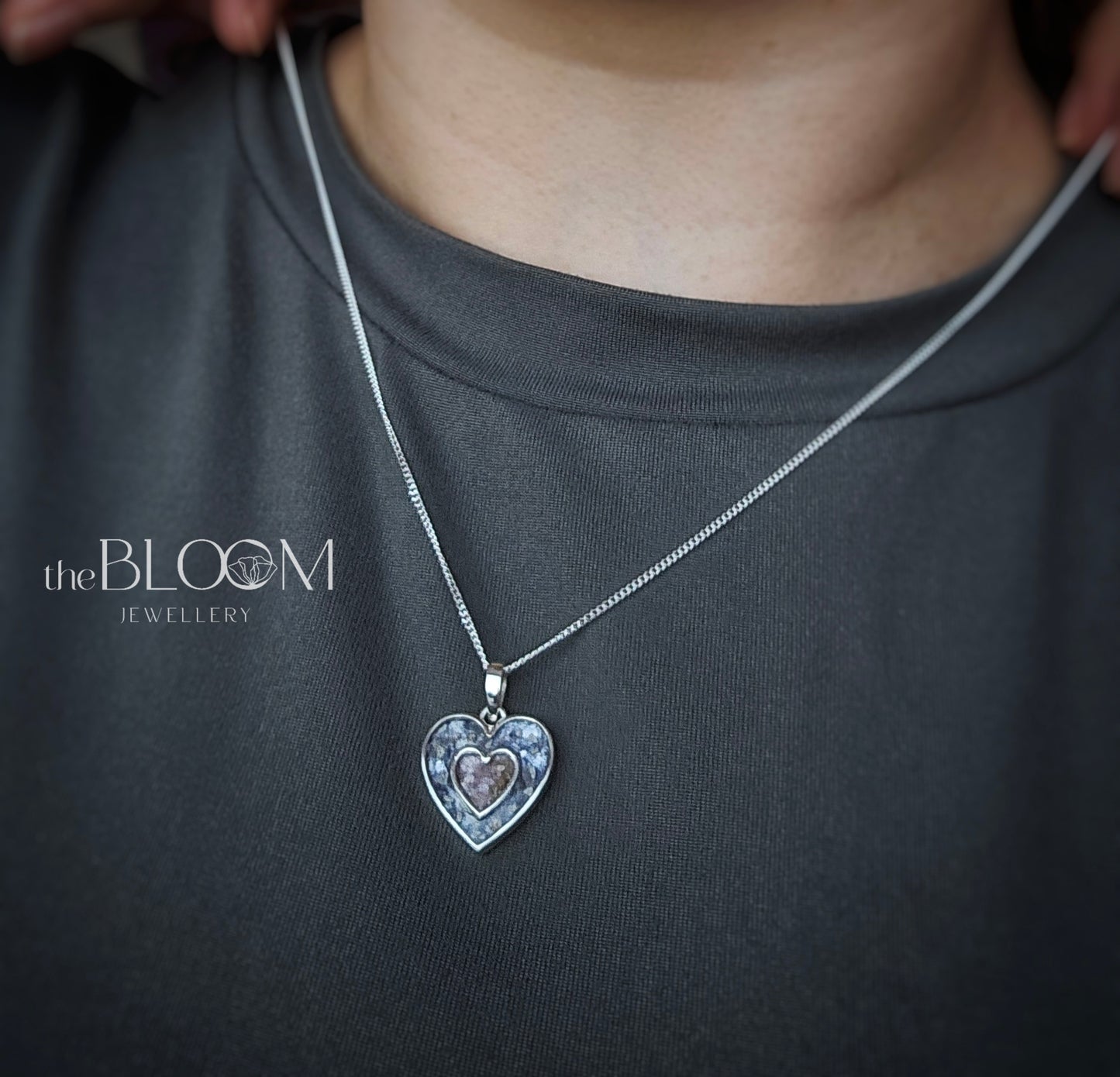 Silver heart-shaped pendant necklace on a person wearing a dark gray shirt, with 'the BLOOM Jewellery' branding.