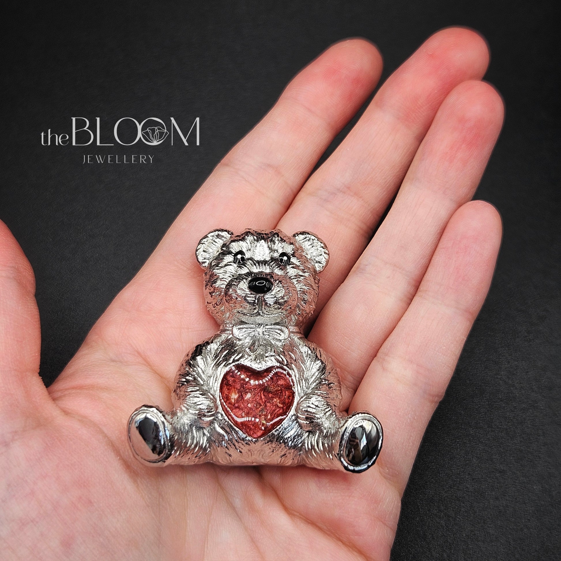 Silver teddy bear pendant with a red heart on a hand against a dark background, branded 'the BLOOM JEWELLERY'.