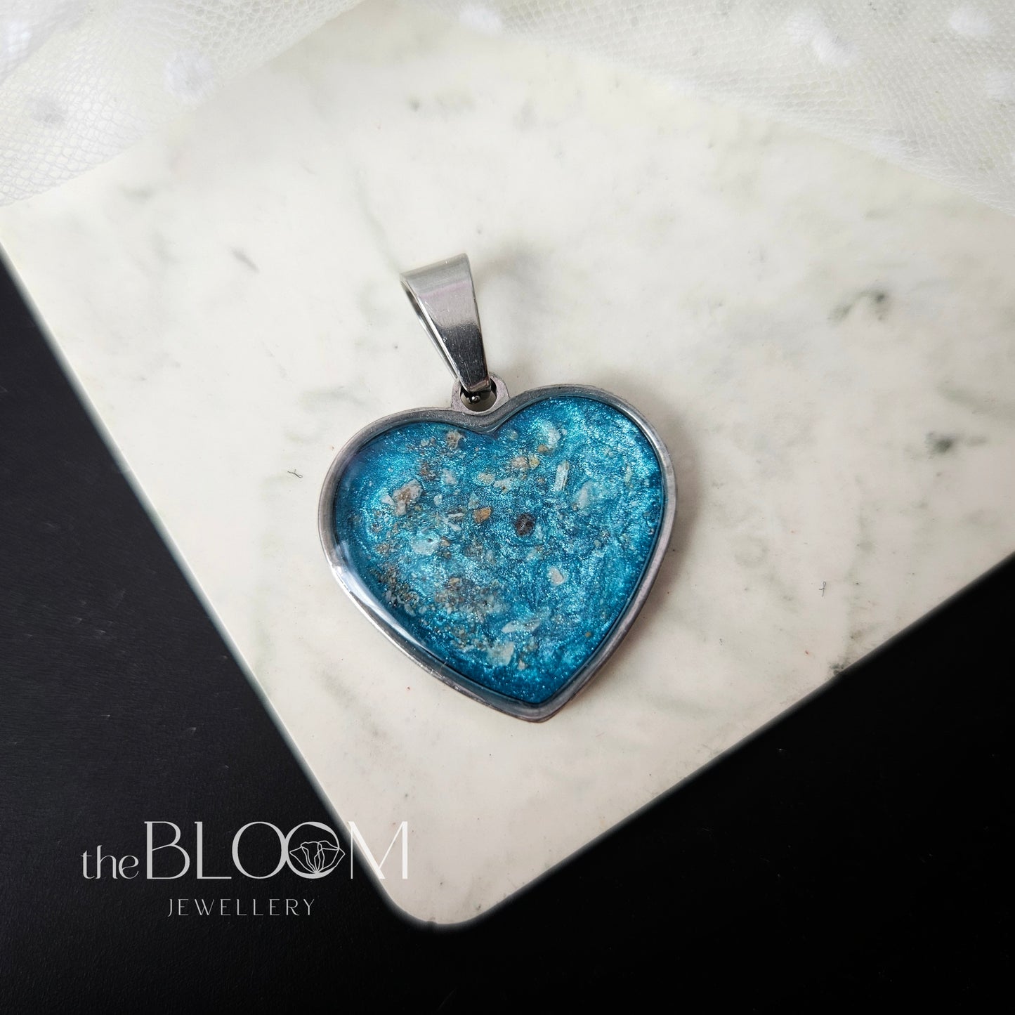 Heart-shaped blue pendant on a marble surface with 'the BLOOM JEWELLERY' branding.