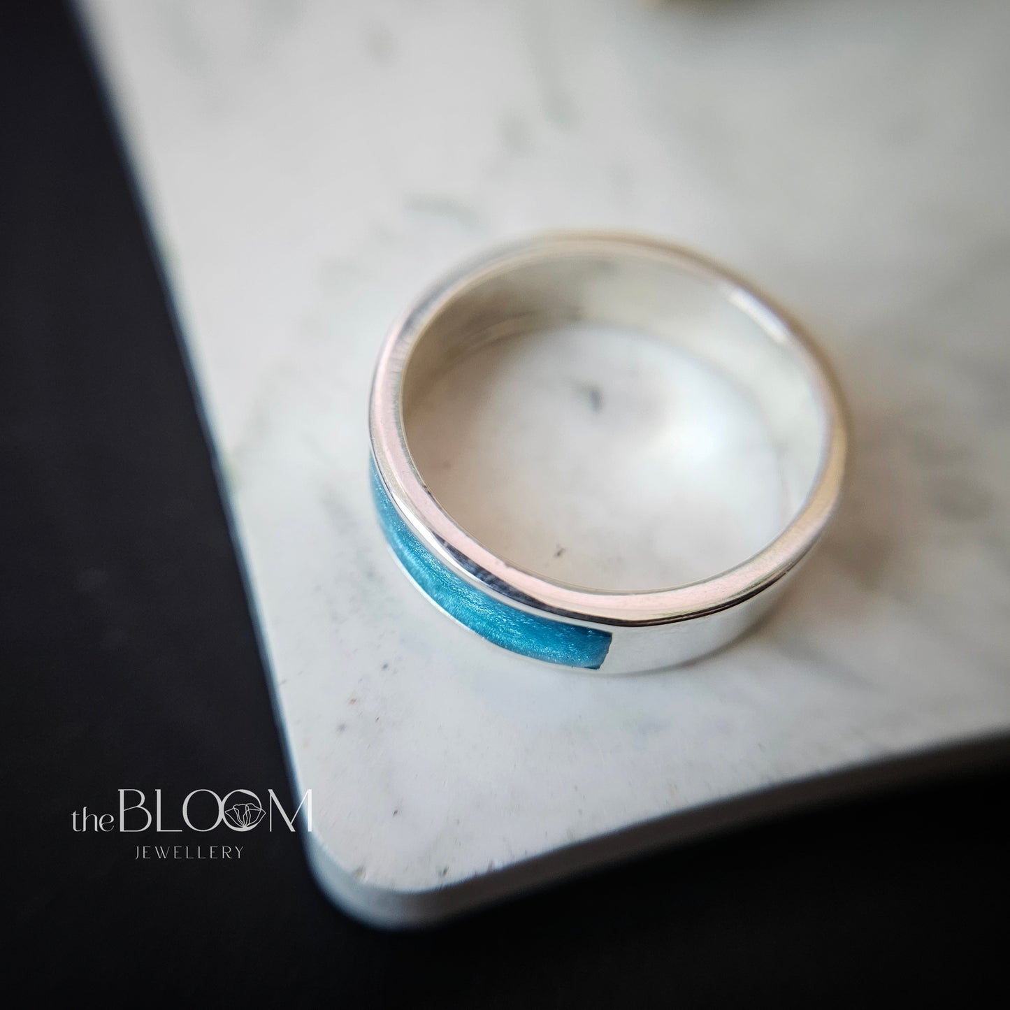 Silver ring with a blue inlay on a marble surface, branded 'theBLOOM JEWELLERY'.