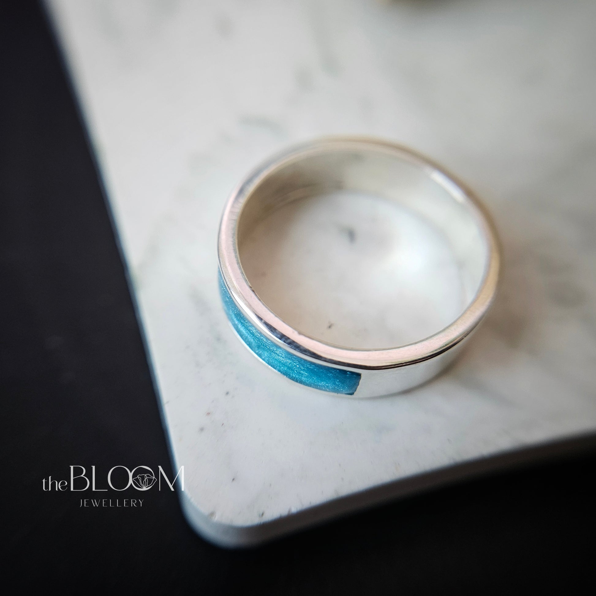 Silver ring with a blue inlay on a marble surface, branded 'theBLOOM JEWELLERY'.
