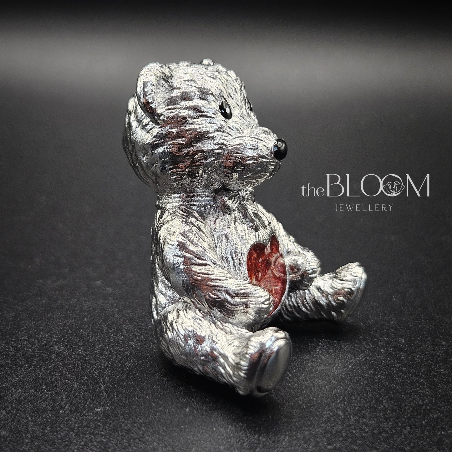 Silver teddy bear figurine with a red heart on a dark background, branded 'the BLOOM JEWELLERY'.