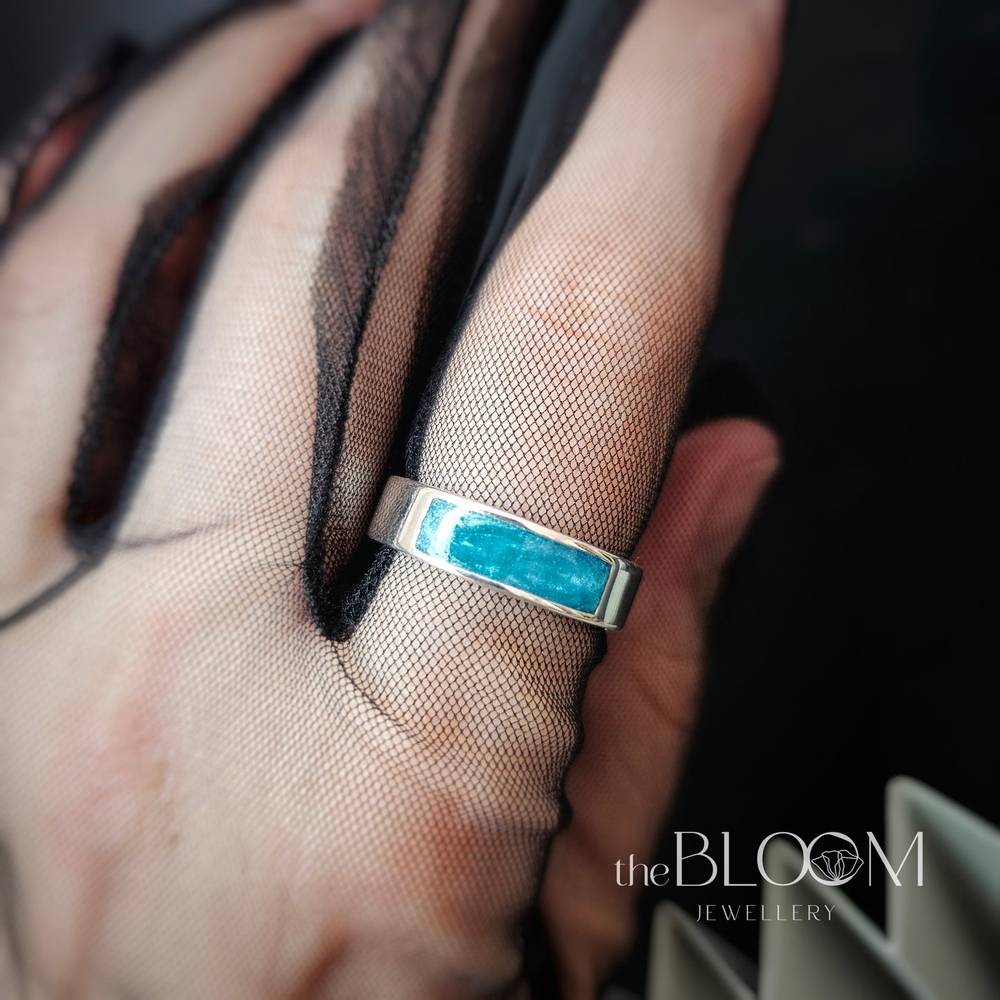 Silver ring with a blue gemstone on a hand, branded 'The Bloom Jewellery'.