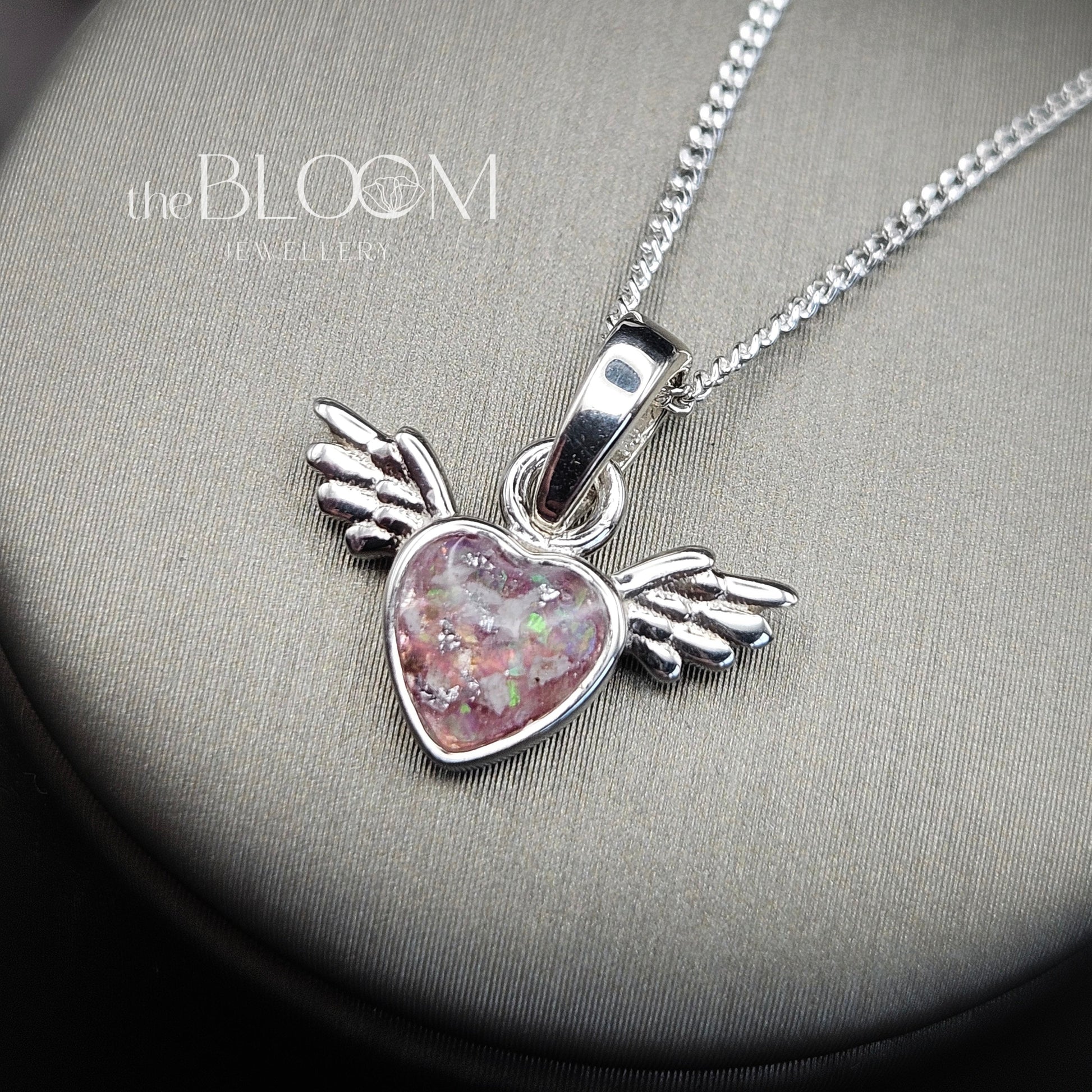 Silver necklace with a heart-shaped pendant featuring wings on a gray surface, branded 'The Bloom'.