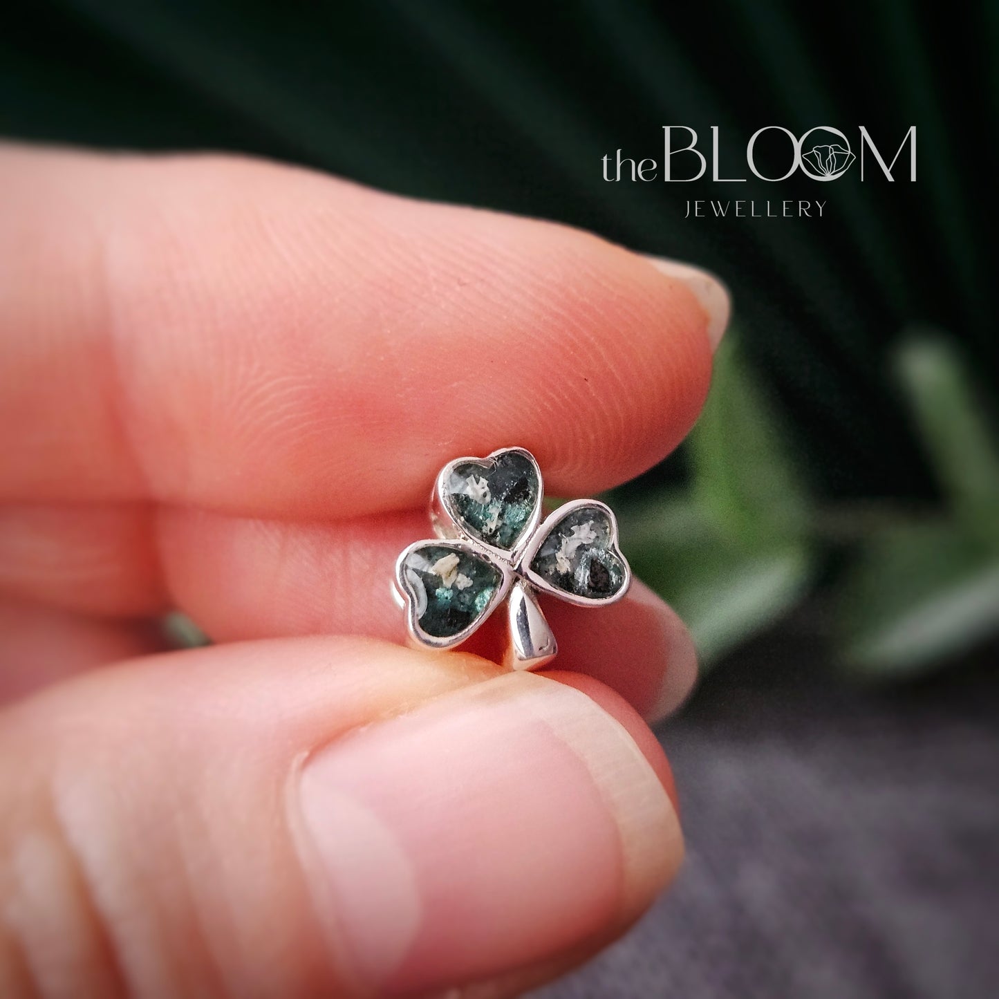 Silver four-leaf clover earring held between fingers with 'the Bloom Jewellery' branding.