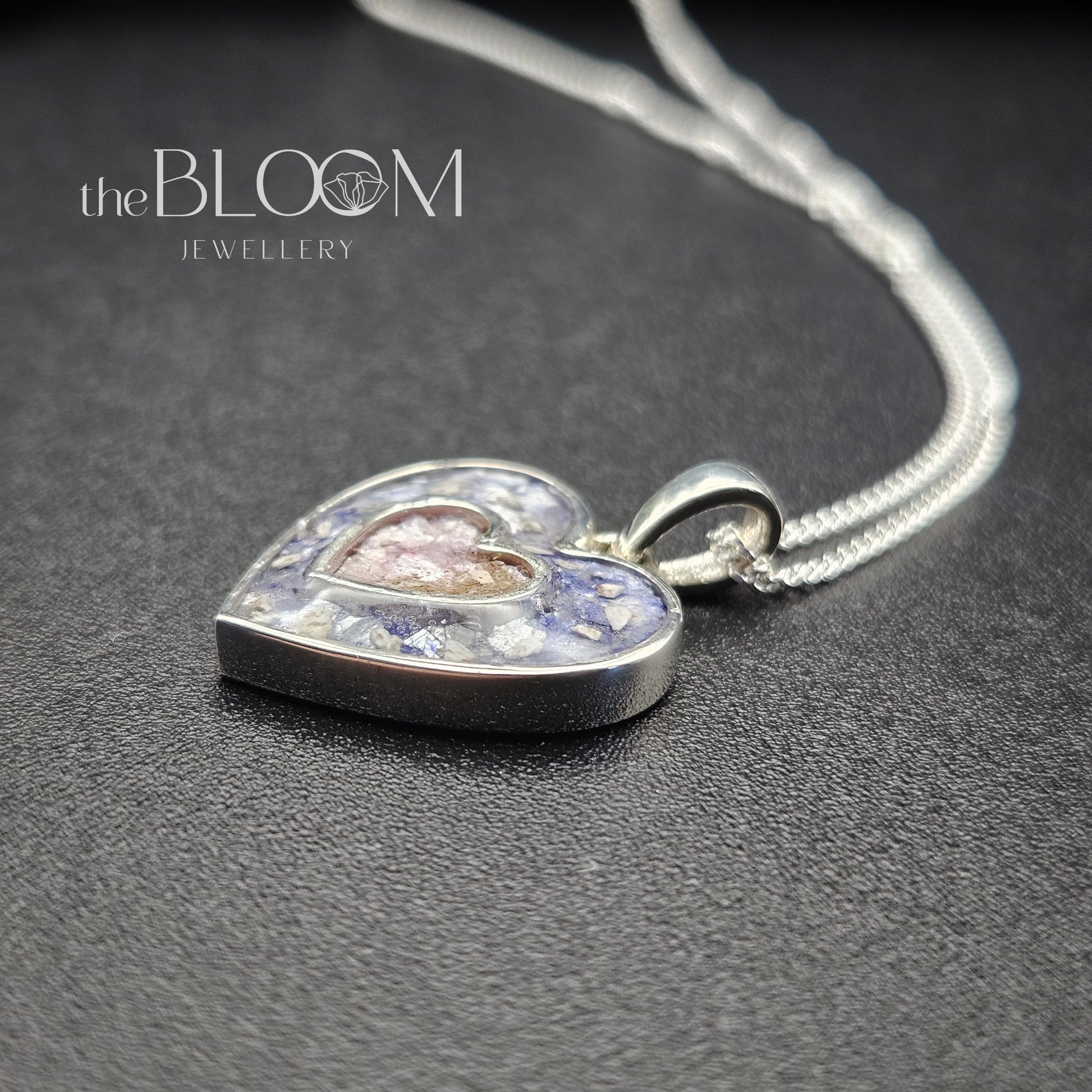 Heart-shaped silver pendant with embedded stones on a dark surface, branded 'the BLOOM JEWELLERY'.