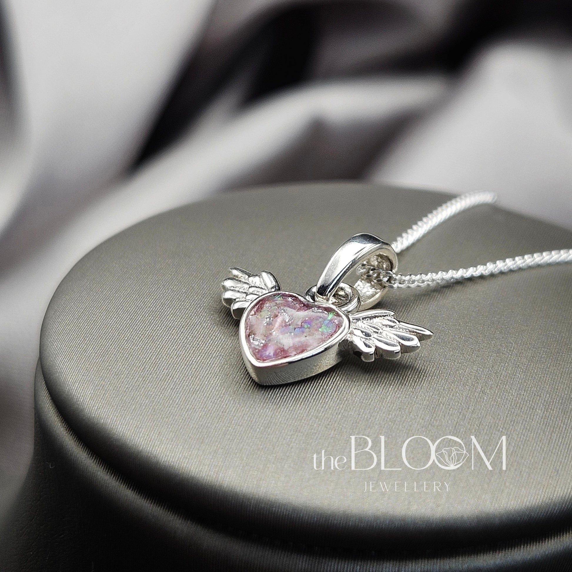 Silver pendant with pink heart and wings on a gray surface, branded 'The Bloom Jewellery'.