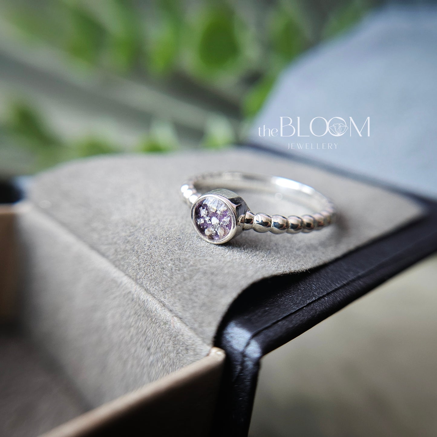 Silver ring with a purple gemstone on a gray surface, branded 'the BLOOM Jewellers'.