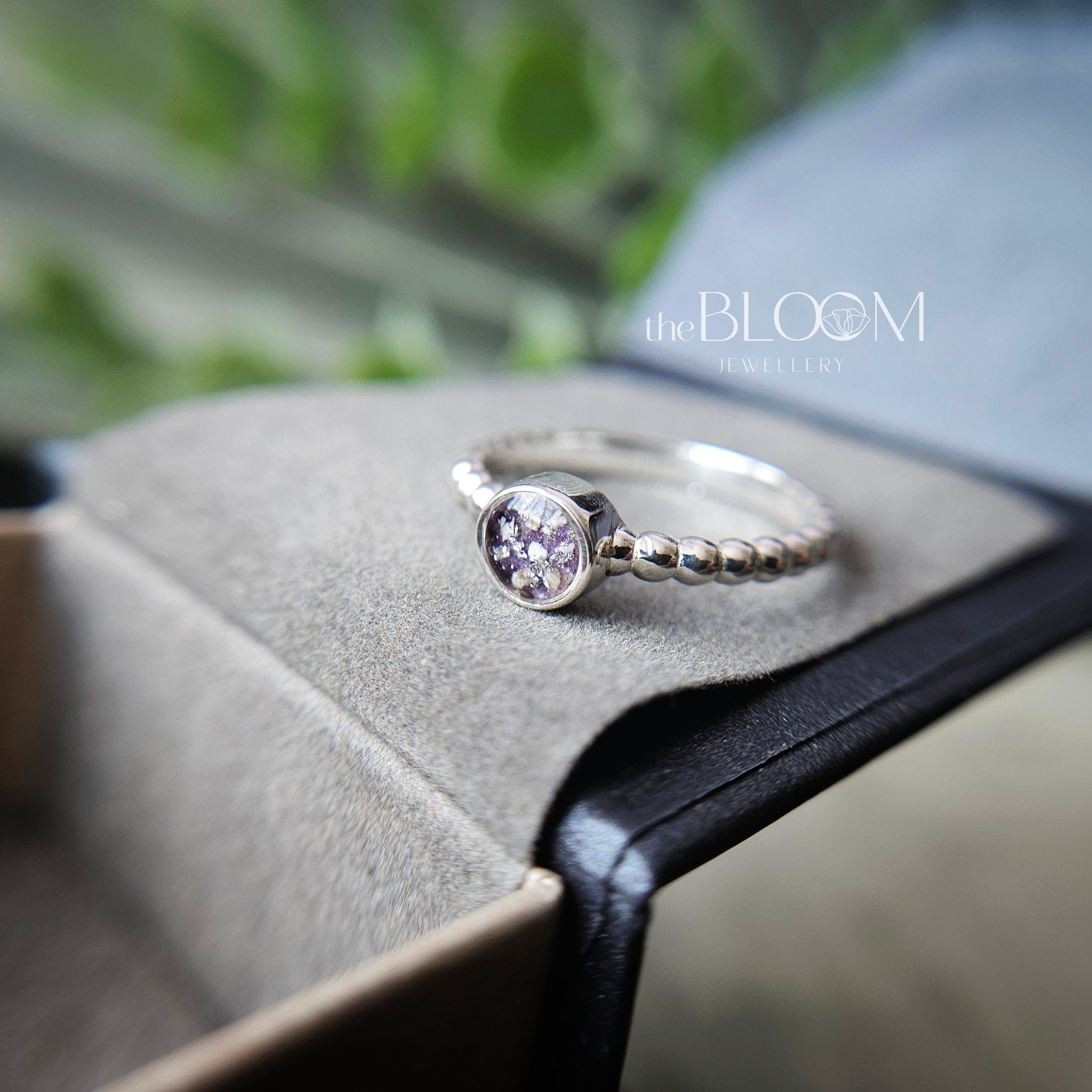Silver ring with a purple gemstone on a gray surface, branded 'the BLOOM Jewellers'.