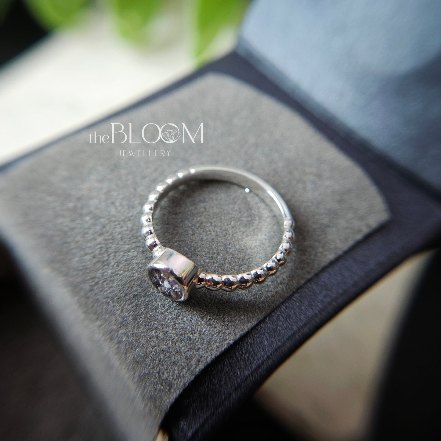 Silver ring with a diamond on a gray surface, branded 'the BLOOM Jewellery'.