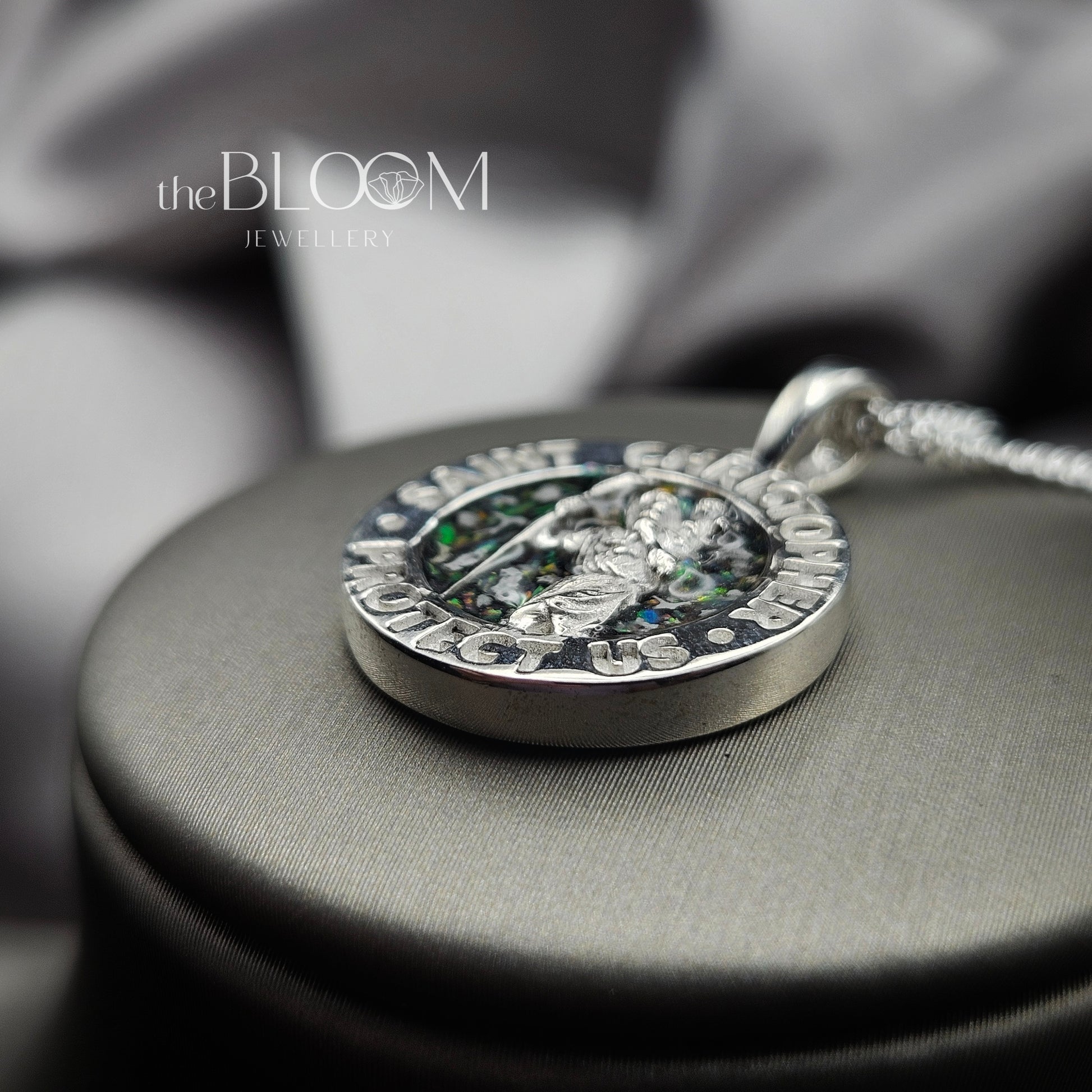 Silver pendant with engraved text on a gray fabric background, branded 'the BLOOM JEWELLERY'.