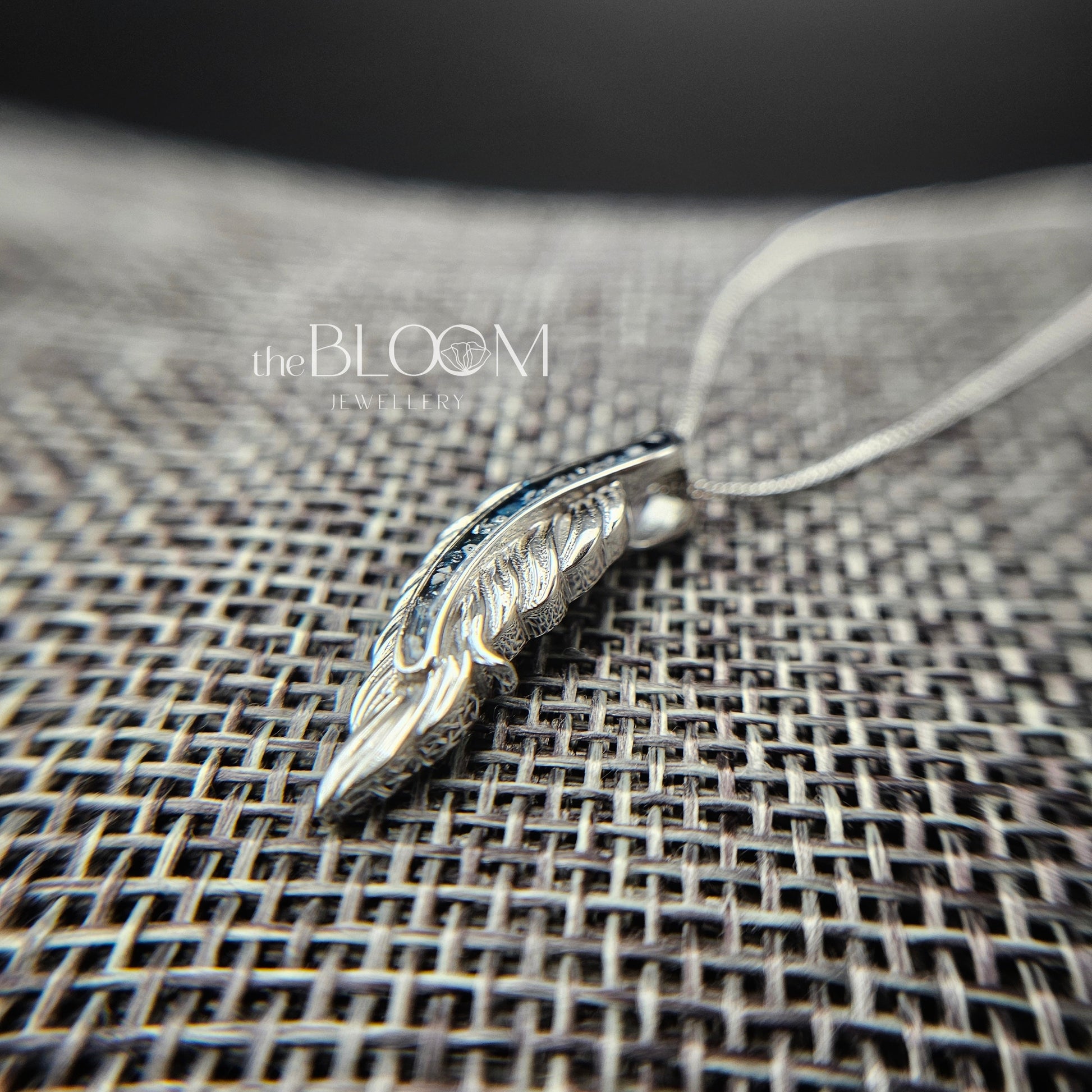 white gold feather pendant on a textured surface with 'the Bloom' branding.