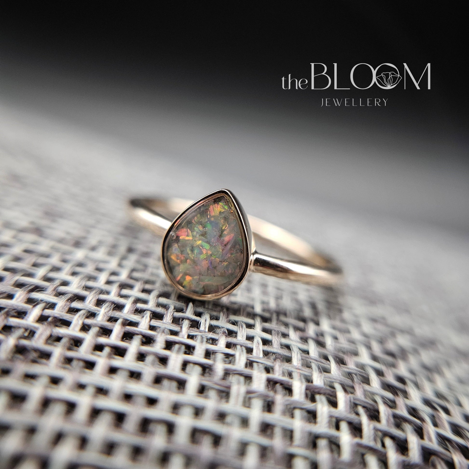 gold ring with a teardrop-shaped opal stone on a textured surface, branded 'the BLOOM'.